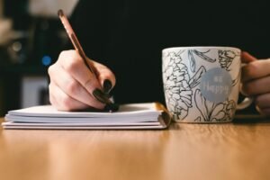 Person writing in a notebook with a floral ceramic mug on a wooden desk.